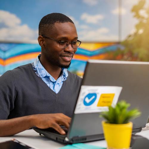 A person of color sitting in a bright room working on a laptop